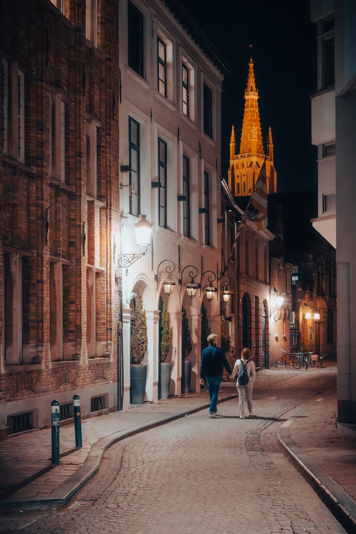 A couple strolls through the charming streets of Bruges at night, with a lit tower in the background.